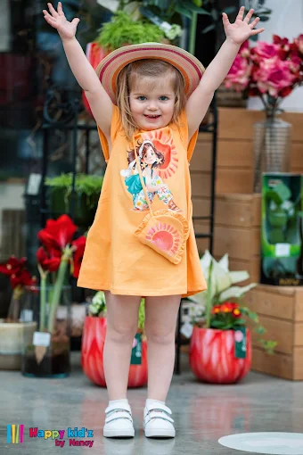 Petite fille souriante en robe orange à motifs et chapeau, un look Happy Kid'z by Nancy.